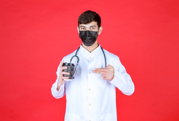 Doctor with stethoscope in black mask holding a black takeaway coffee cup. 