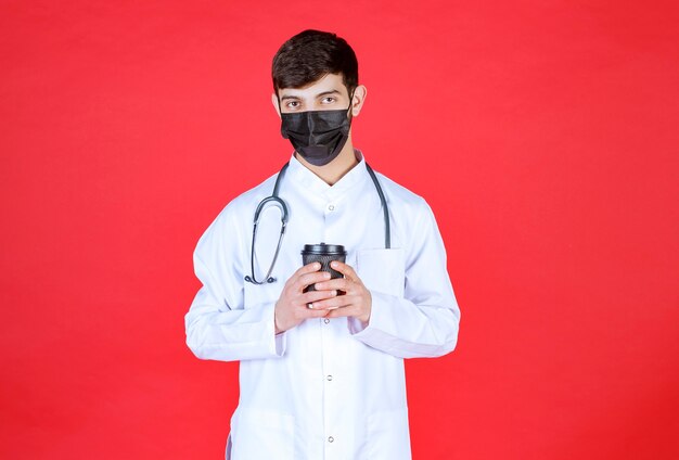 黒い持ち帰り用のコーヒーカップを持っている黒いマスクの聴診器を持つ医師。