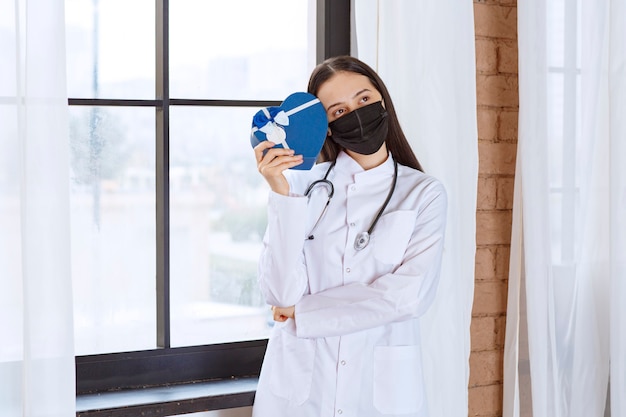 Доктор со стетоскопом и черной маской держит синюю подарочную коробку в форме сердца и выглядит смущенным и неуверенным.