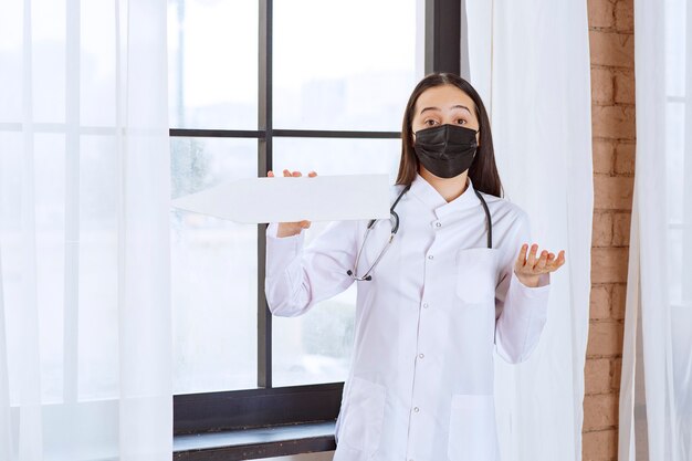 Doctor with stethoscope and black mask holding a white arrow pointing left and looks confused. 