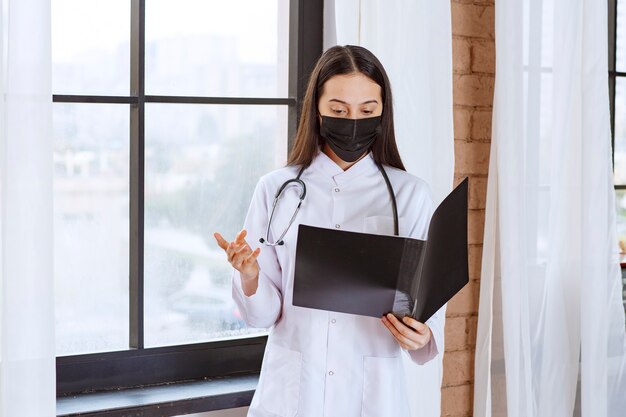 Doctor with stethoscope and black mask standing next to the window and holding a black history folder of the patients, checking it and looks terrified. 