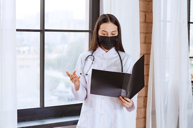 聴診器と黒いマスクを持った医師が窓の隣に立って、患者の黒い履歴フォルダを持って、それをチェックして、おびえているように見えます。
