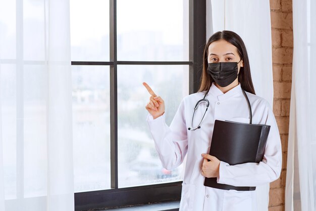 Doctor with stethoscope and black mask standing next to the window and holding a black history folder of the patients while pointing at somewhere. 