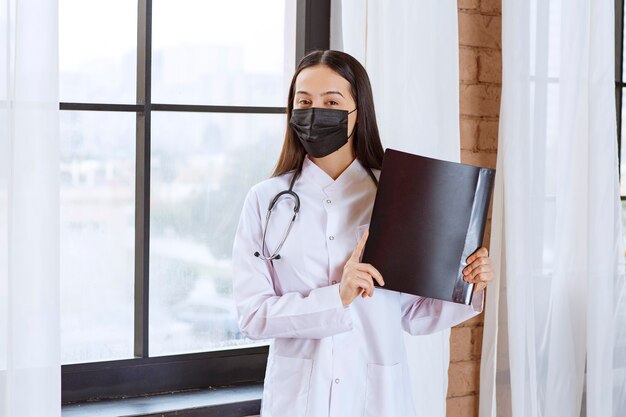 Doctor with stethoscope and black mask standing next to the window and holding a black history folder of the patients. 