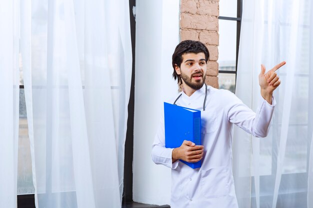 Doctor with a stethoscope holding a blue reporting folder and pointing at someone around. 