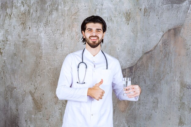 순수한 물 한 잔을 들고 긍정적인 손 기호를 보여주는 청진기를 가진 의사