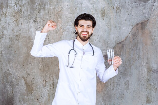 Doctor with a stethoscope holding a glass of pure water and showing positive hand sign. 