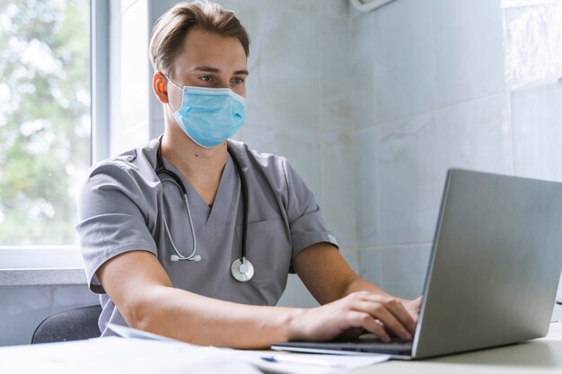 Doctor with stethoscope and medical mask working on laptop