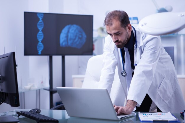 Доктор работает на ноутбуке с изображением мозга на заднем плане. Врач-кардиолог за работой.