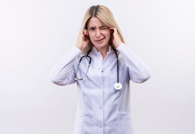 Доктор молодая блондинка со стетоскопом и медицинским халатом с закрытыми ушами на изолированной белой стене