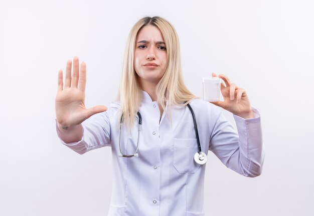 聴診器と空の缶を保持している医療用ガウンを身に着けている医者の若いブロンドの女の子は、孤立した白い壁に停止ジェスチャーを示すことができます