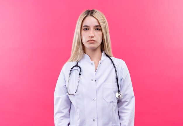 孤立したピンクの壁に医療用ガウンで聴診器を身に着けている医者若いブロンドの女の子