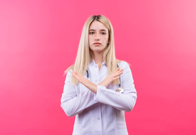 Medico giovane ragazza bionda che indossa uno stetoscopio in abito medico che mostra il gesto no sulla parete rosa isolata