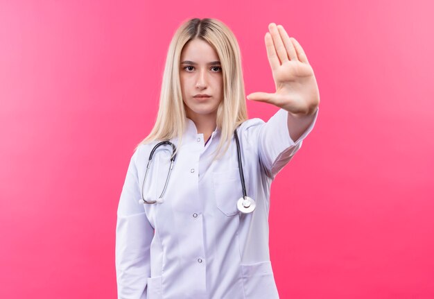 孤立したピンクの壁に停止ジェスチャーを示す医療ガウンで聴診器を身に着けている医者若いブロンドの女の子