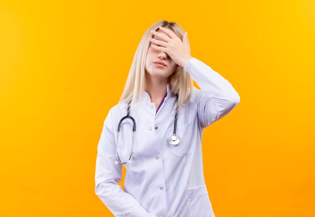 Doctor young girl wearing stethoscope in medical gown covered eyes with hand on isolated yellow wall