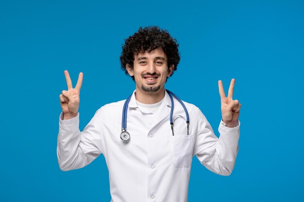 Doctors day curly brunette cute guy in lab coat showing peace gesture