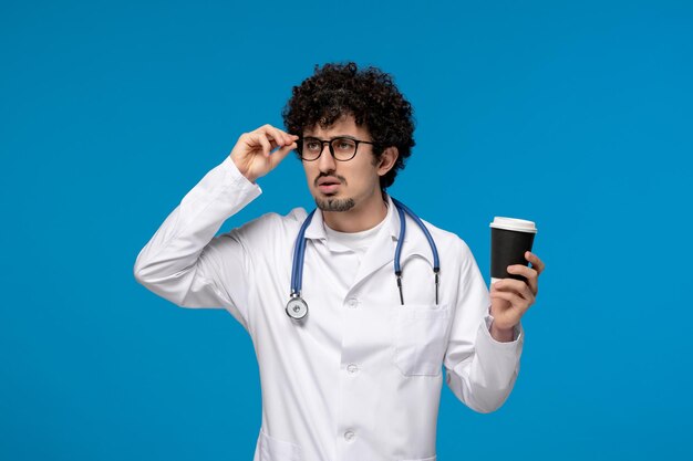 Doctors day curly brunette cute guy in medical uniform holding a paper cup and thinking