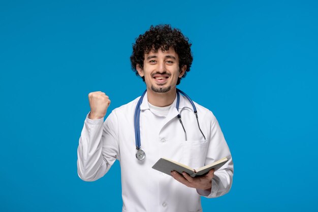 Doctors day curly handsome cute guy in medical uniform holding fist up and a book
