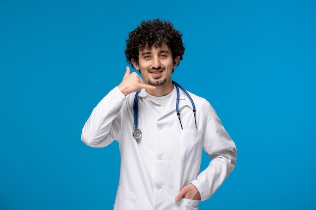 Doctors day curly handsome cute guy in medical uniform making phone call gesture