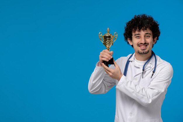 Doctors day curly handsome cute guy in medical uniform showing a golden trophy