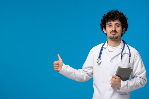 Doctors day curly handsome cute guy in medical uniform showing good gesture and holding book
