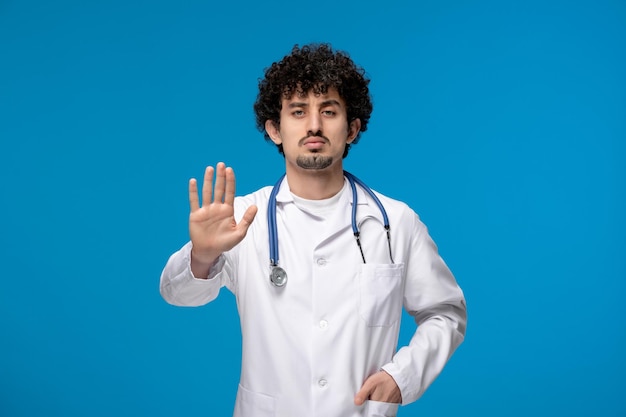 Doctors day curly handsome cute guy in medical uniform showing stop sign