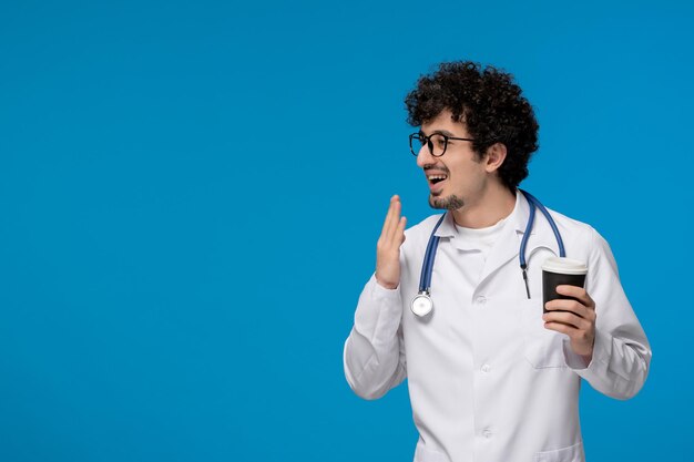 Doctors day curly handsome cute guy in medical uniform wearing glasses with papercup