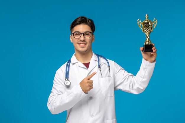 Doctors day cute young brunette guy in lab coat wearing glasses smiling and holding golden trophy