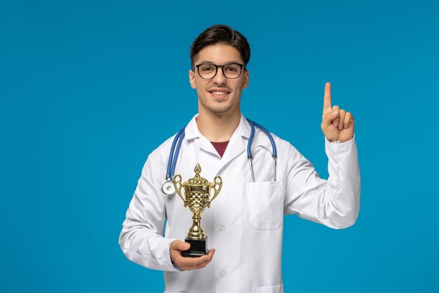 Doctors day cute young brunette guy in lab coat wearing glasses with phonendoscope and trophy