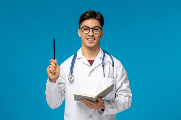 Doctors day cute young handsome man in lab coat and glasses excitingly holding pen and book