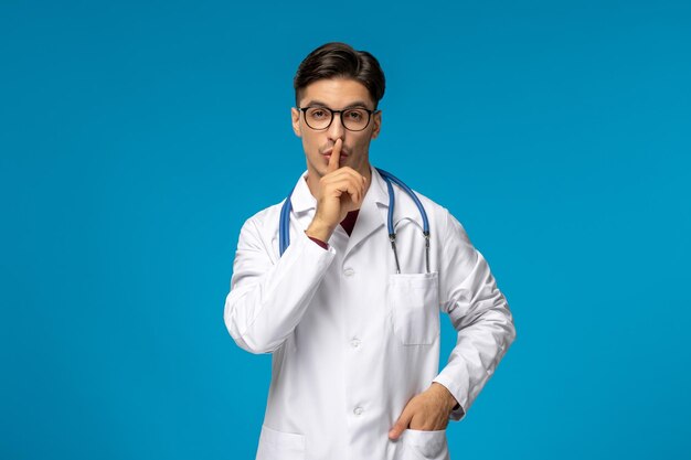 Doctors day handsome brunette cute guy in medical gown showing silence gesture