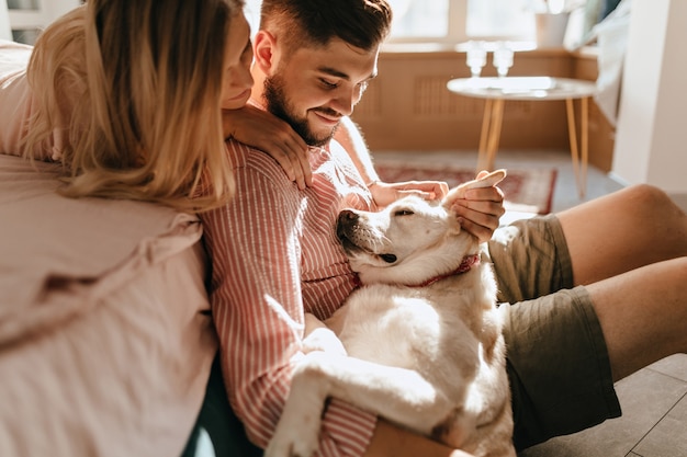 Free photo dog lies on legs of owner. man in pink shirt and his beloved woman admire their white pet.