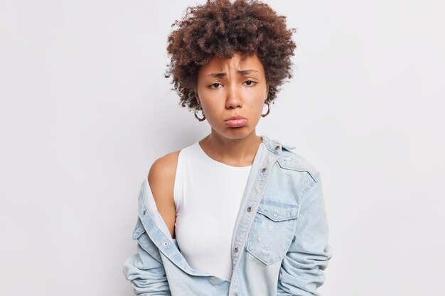 Doleful Afro American woman purses lower lip frowns face from something bad makes unhappy grimace being offended dressed in stylish clothes poses against white wall