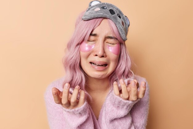 Doleful dejected young woman with eastern appearance gestures from despair dressed in casual jumper and sleepmask on forehead applies hydrogel patches under eyes isolated over beige background