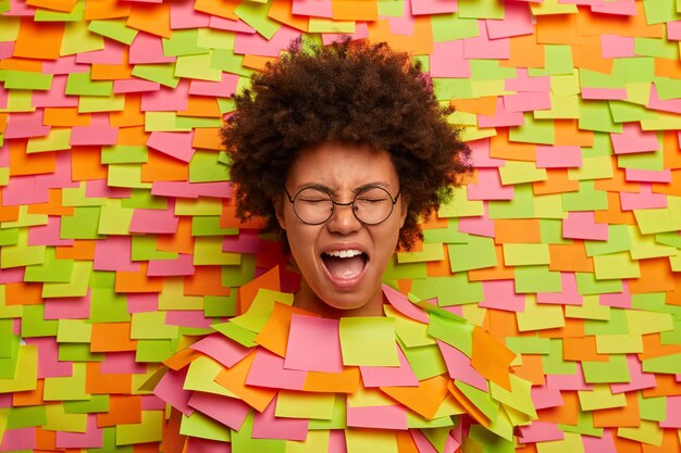 Doleful distressed ethnic woman cries from despair, keeps mouth widely opened, suffers because of breakdown, whins loudly, screams and complains on something, poses through paper wall, stickers around