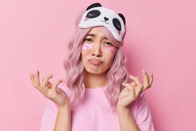Doleful pink haired woman has eastern appearance spreads hands feels upset applies hydrogel patches under eyes for skin moisturising wears sleepmask and casual t shirt isolated over pink wall