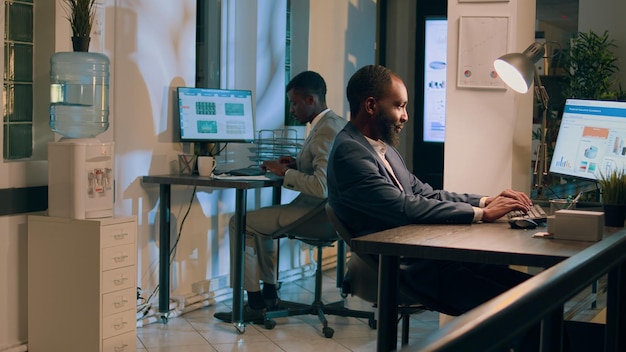 Free photo dolly out shot of african american employees sitting at desks in office, imputing data on computers overnight. coworkers in modern relaxed workspace solving tasks during nightshift