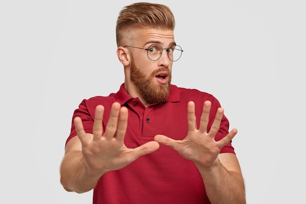 Don`t bother me please! Surprised emotional ginger bearded young guy makes stop gesture, stratches hands, feels displeased, wears casual red t-shirt, isolated over white wall.