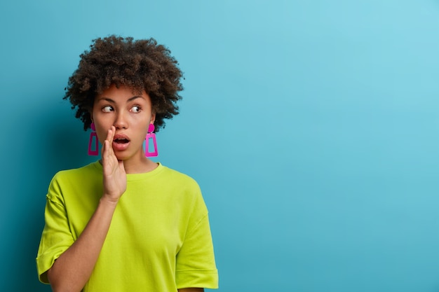 Dont tell anyone! Shocked talkative young woman keeps palm near mouth, looks aside, tells private infomation and spreads rumors, impressed by surprised sudden news, isolated on blue wall