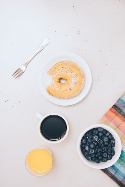 Donut with bite missing; juice; coffee cup and blue berries on white backdrop