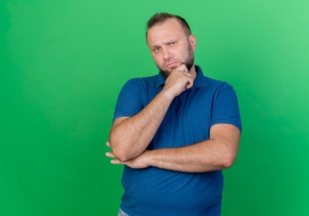 Doubtful adult slavic man putting hands under elbow and on chin  isolated on green wall with copy space