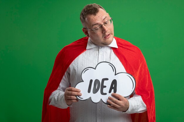 Doubtful adult slavic superhero man in red cape wearing glasses holding and looking at idea bubble isolated on green wall with copy space