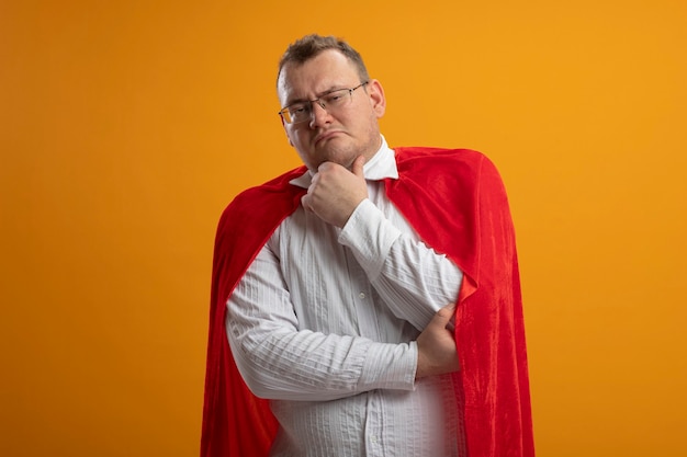 Doubtful adult superhero man in red cape wearing glasses looking at front touching chin isolated on orange wall