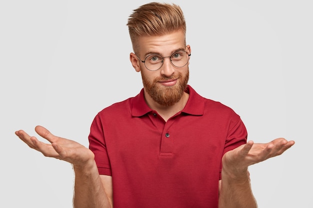 Бесплатное фото Сомнительно привлекательный бородатый молодой мужчина с рыжими волосами, густой бородой и усами, пожимает плечами, сомневается, что купить, привлекательно выглядит, позирует на фоне белой стены. концепция неуверенности