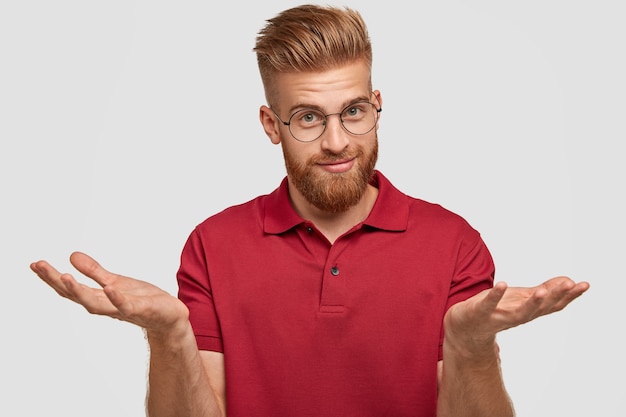 Free photo doubtful attractive bearded young male with ginger hair, thick beard and mustache, shruggs shoulders, doubts what to buy, has appealing look, poses against white wall. hesitation concept