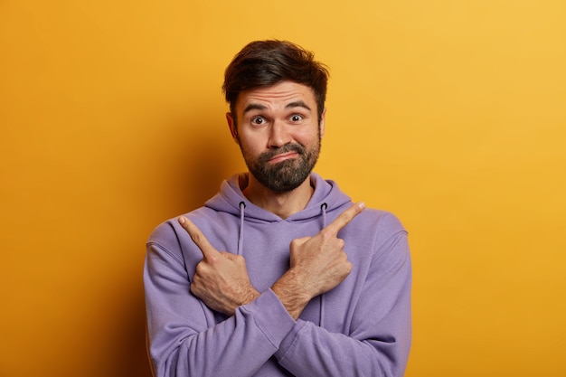 Doubtful bearded guy crosses hands and points index fingers at opposite sides, has difficulty in choosing something, purses lips with hesitation, wears hoodie, isolated on yellow wall.