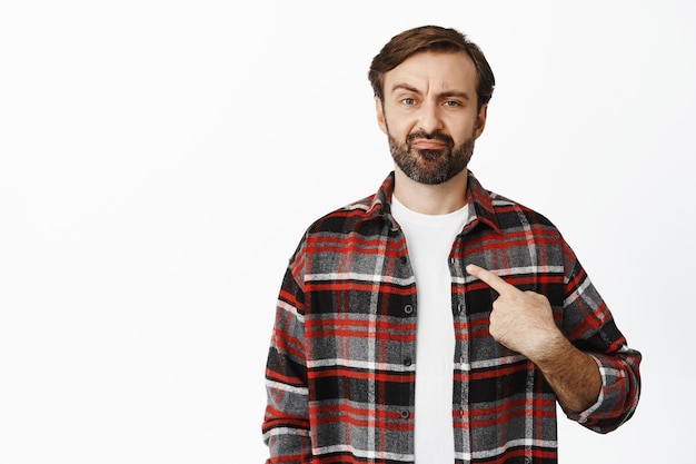 Doubtful bearded man pointing at himself looking hesitant and suspicious frowning and grimacing standing over white background