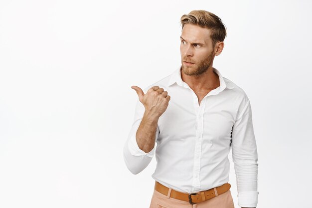 Doubtful blond man pointing and looking left with concerned puzzled face expression having doubts white background