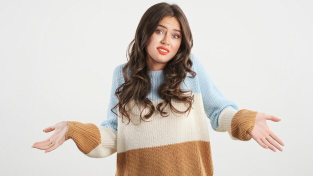 Free photo doubtful brunette girl in knitted sweater throwing up hands guiltily looking in camera over white background i dont know expression