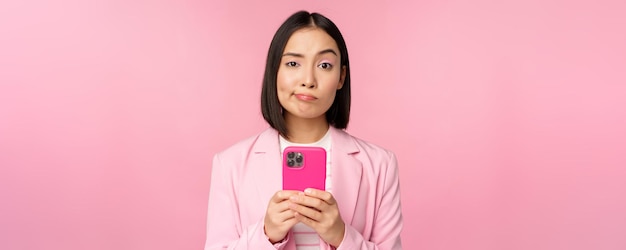 Сомнительная деловая женщина в костюме, держащая смартфон, гримасничает со скептическим выражением лица, стоя на розовом фоне